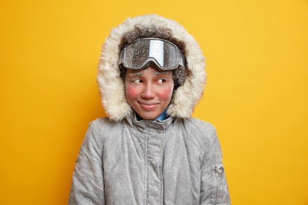 Мечтательная зимняя девушка с красным замороженным лицом наслаждается отдыхом на горном курорте в холодный, добрый снежный день, покрытый снежинками, одетая в теплую куртку с капюшоном, носит лыжные очки, любит экстремальный спорт.