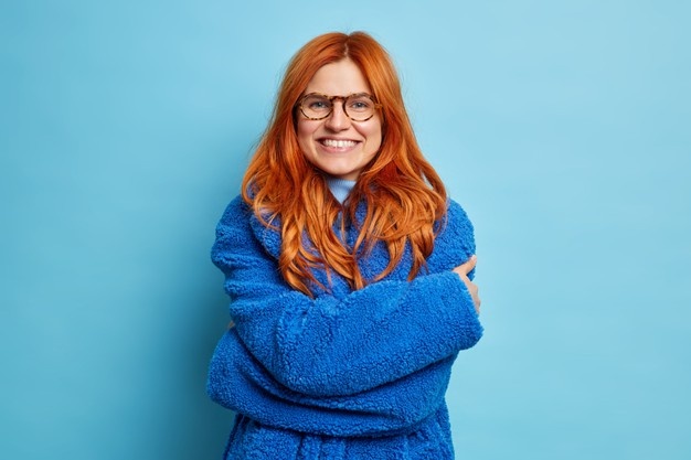 На фото улыбающаяся рыжая европейка обнимает себя и показывает белые зубы, одетая в мягкую ткань пальто, чувствуя тепло в хорошем настроении.